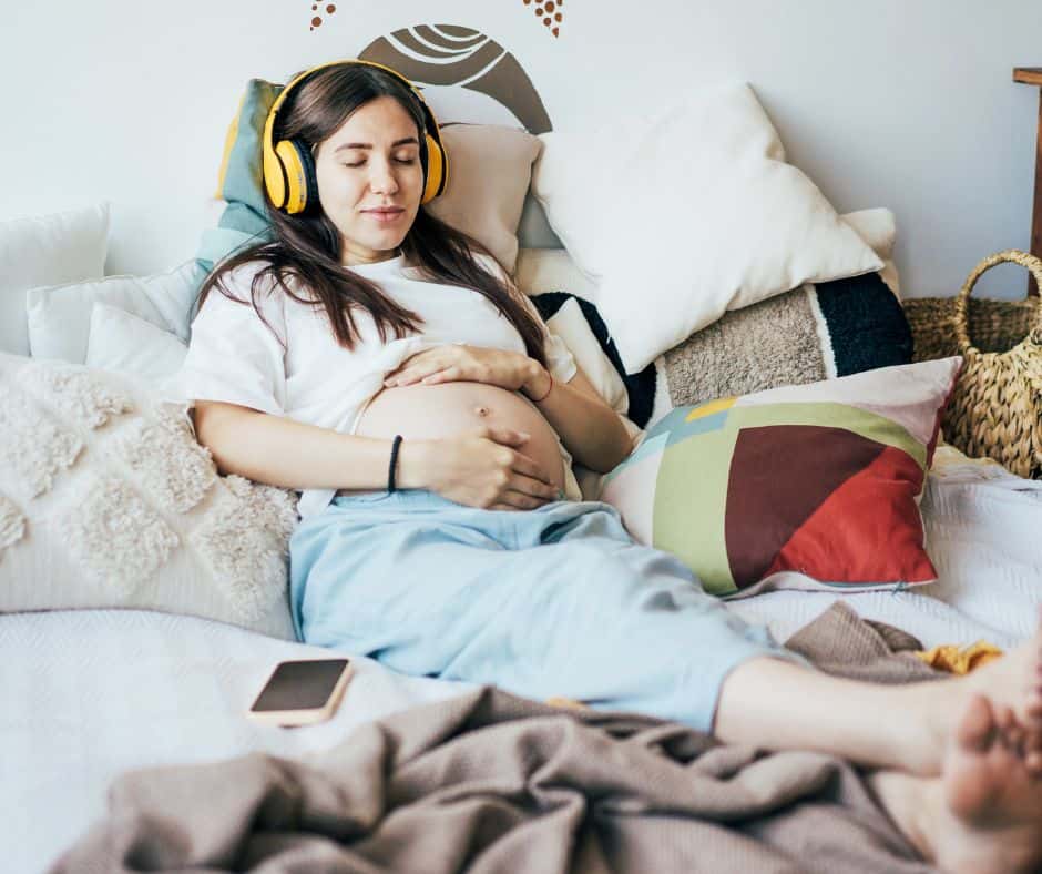 pregnant woman resting in bed with headphones and cradling her belly with her hands