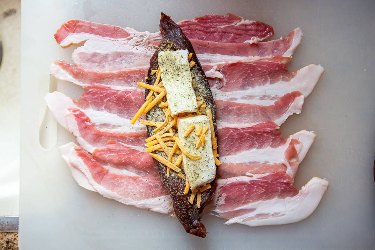 cheese laying on butterflied venison tenderloin, sprinkled with seasoning, with bacon.